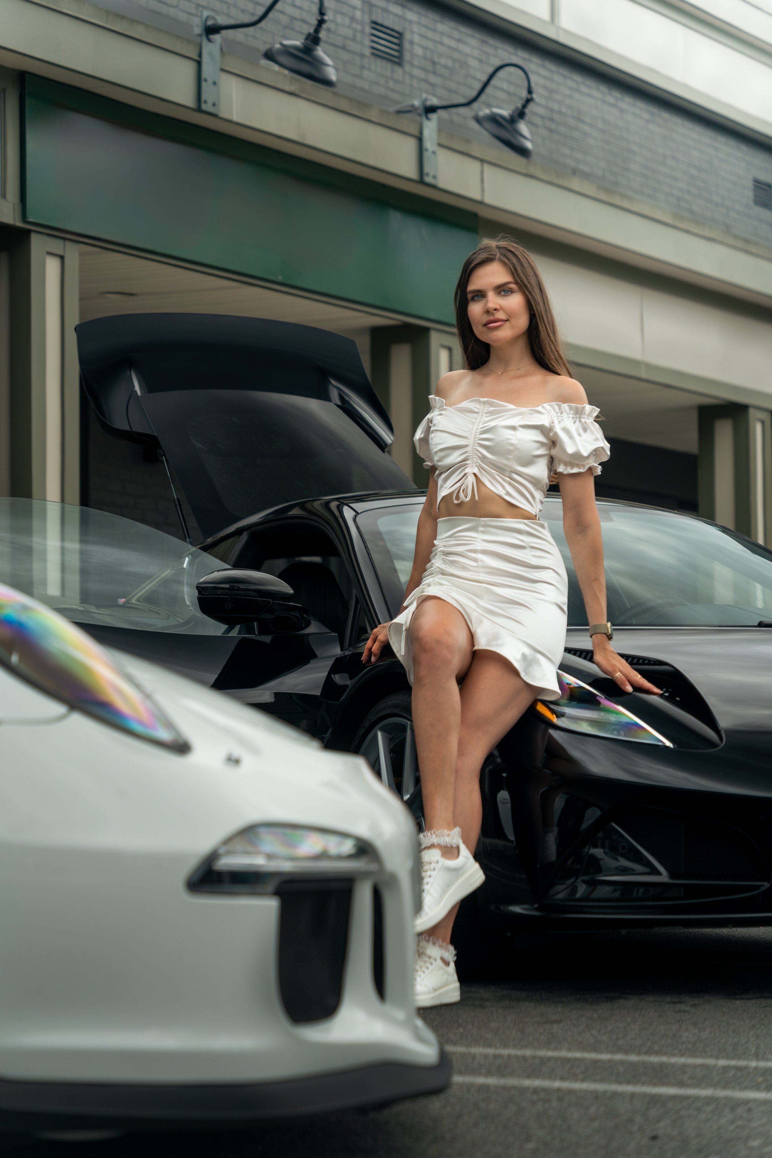 A model in a white dress and sneakers sitting on the hood of a black Lotus Emira with the hood open, in a parking lot near a building at Impavido Exotics