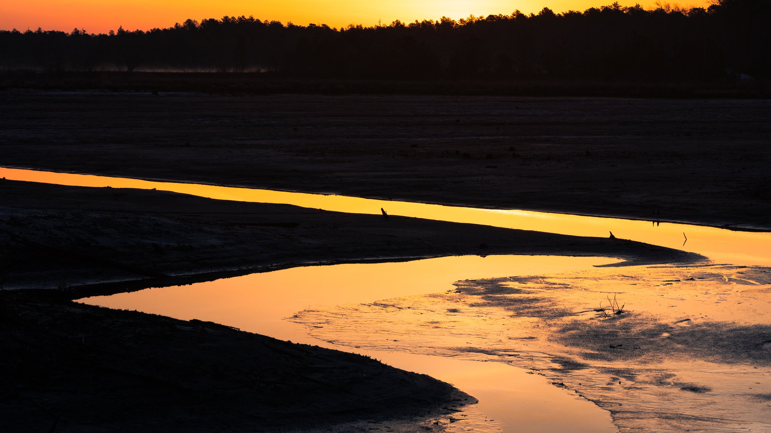Sunset over a river with calm, reflective water and silhouettes of trees on the horizon.