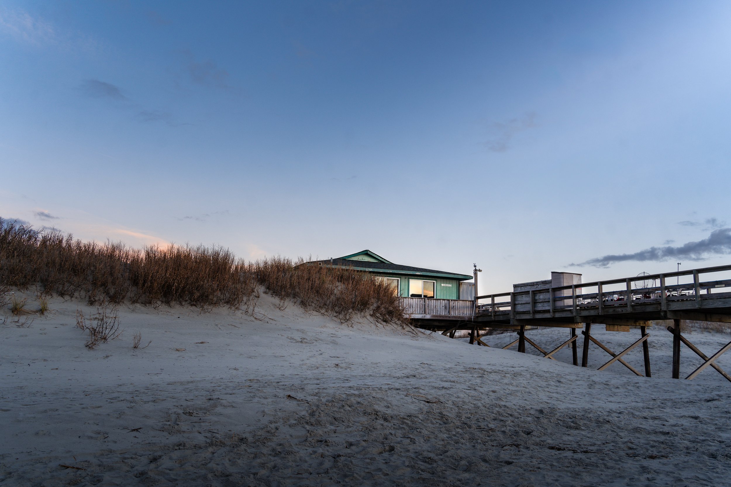 A beach scene with sand dunes covered in sparse vegetation, a green house on stilts, and a wooden walkway under a clear blue sky.
