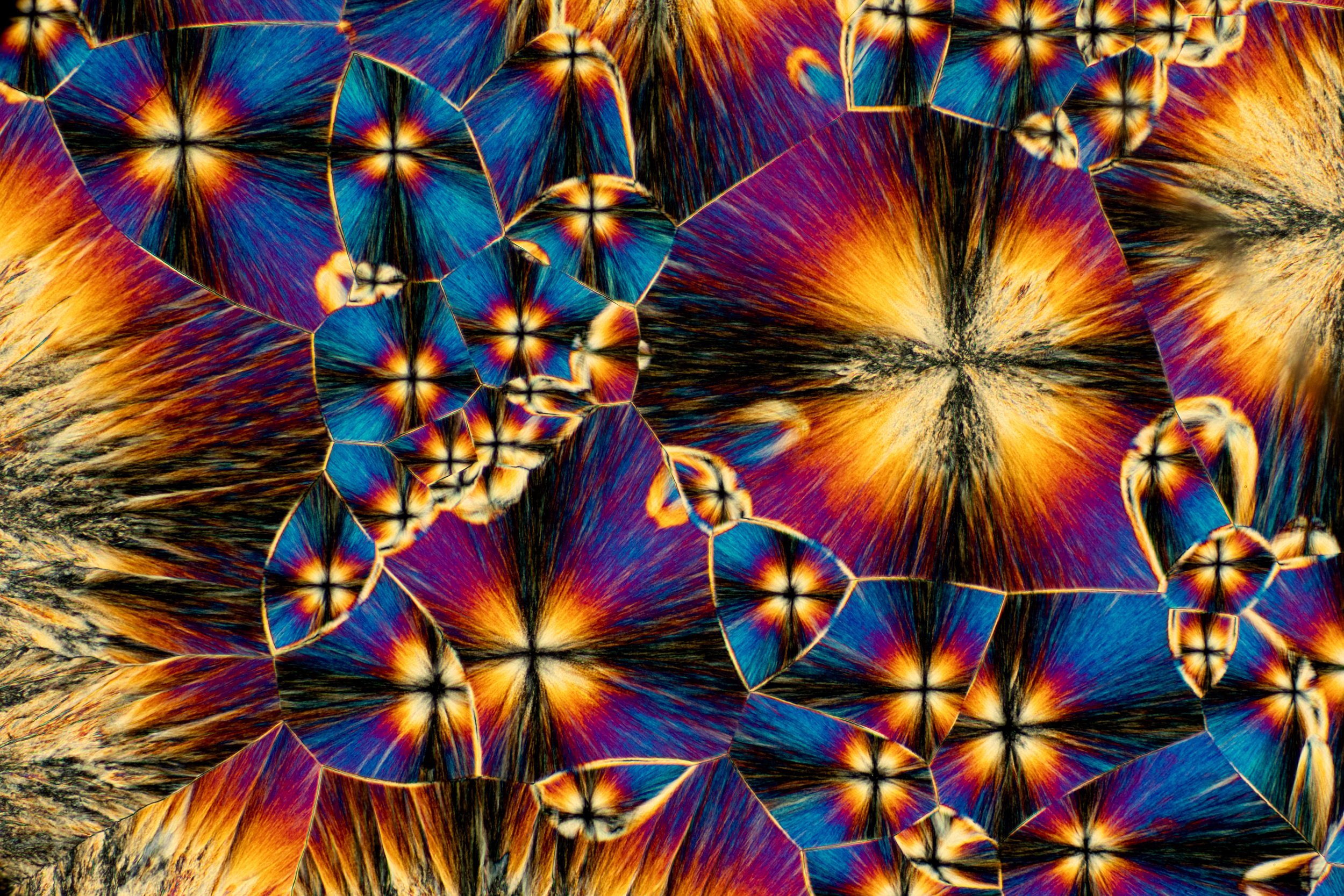 Microscopic cross-polarization of a mineral or crystal with vibrant blue, orange, yellow, and black patterns, showing crystalline structures.