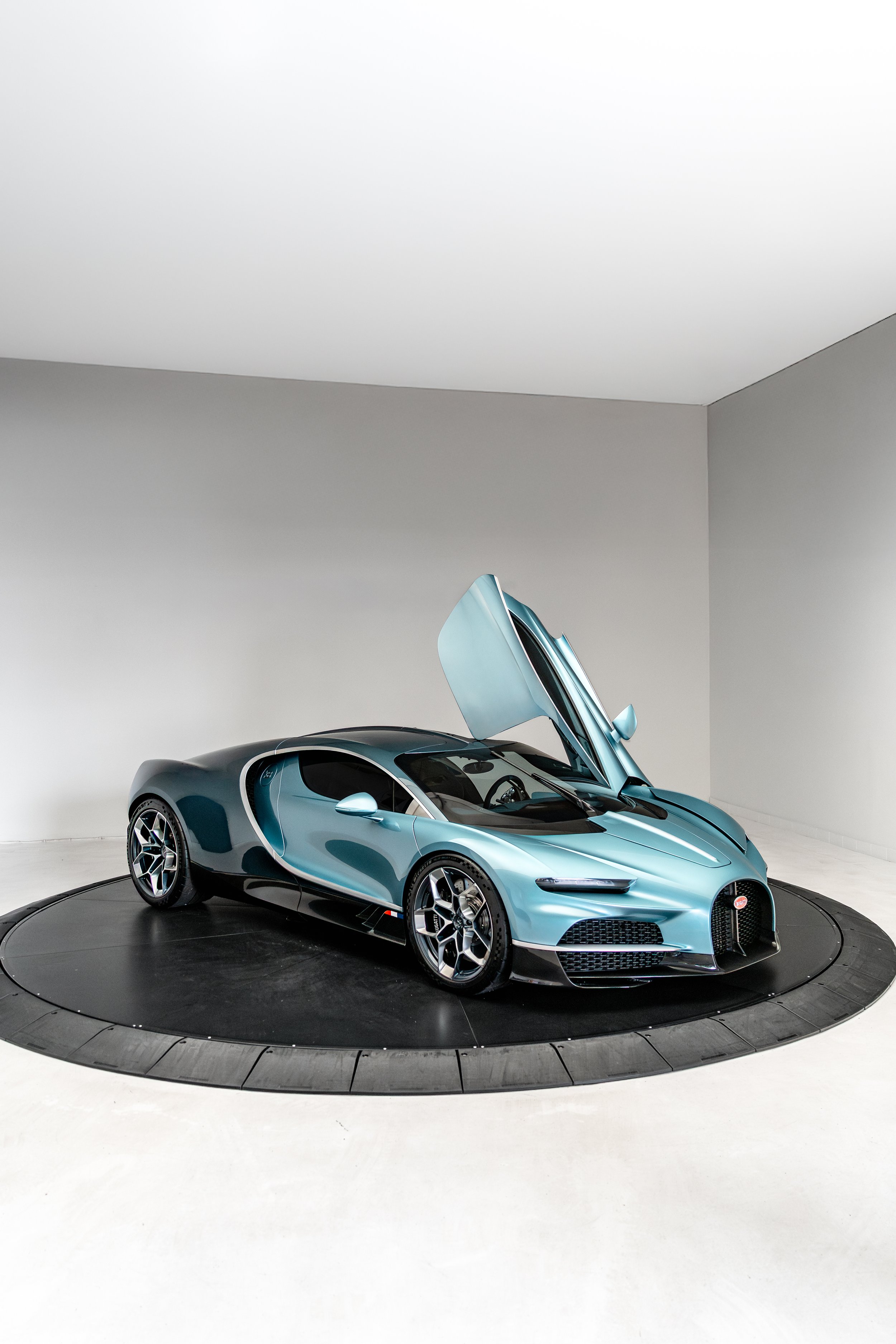 Bugatti Tourbillon with gullwing doors open, displayed on a black circular platform in a white room.