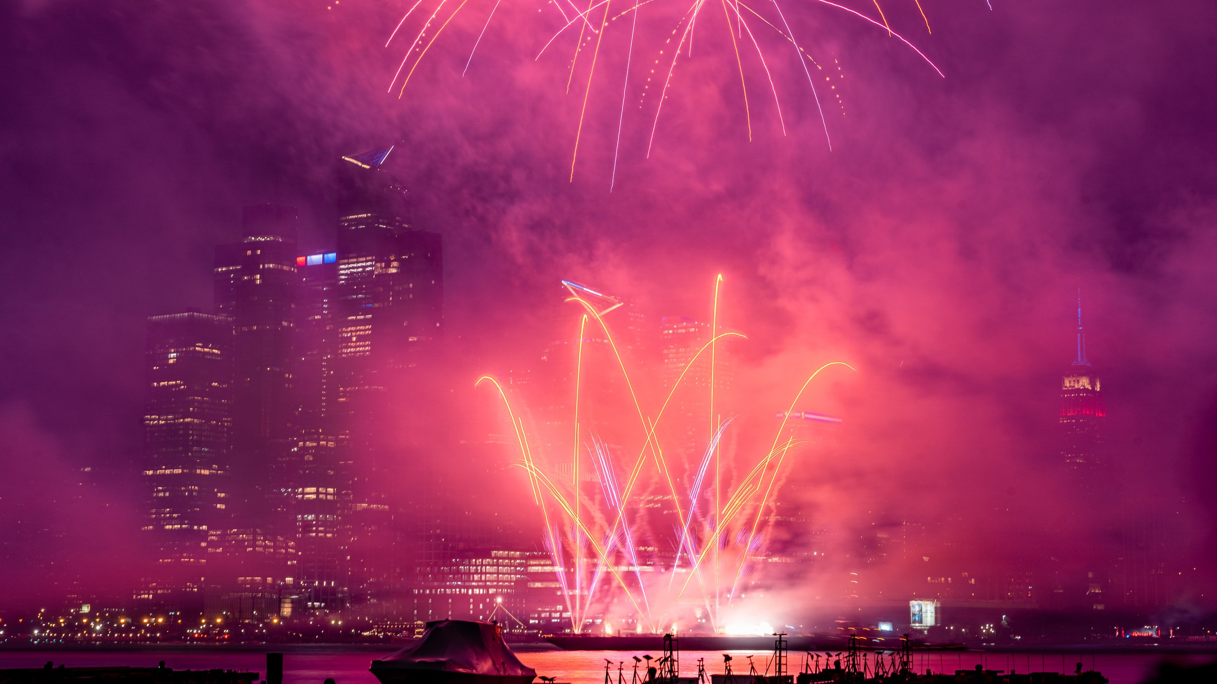 Fireworks display over a city skyline at night with tall buildings and light reflections on the water at the Macy's Fourth of July Firework Show in NYc