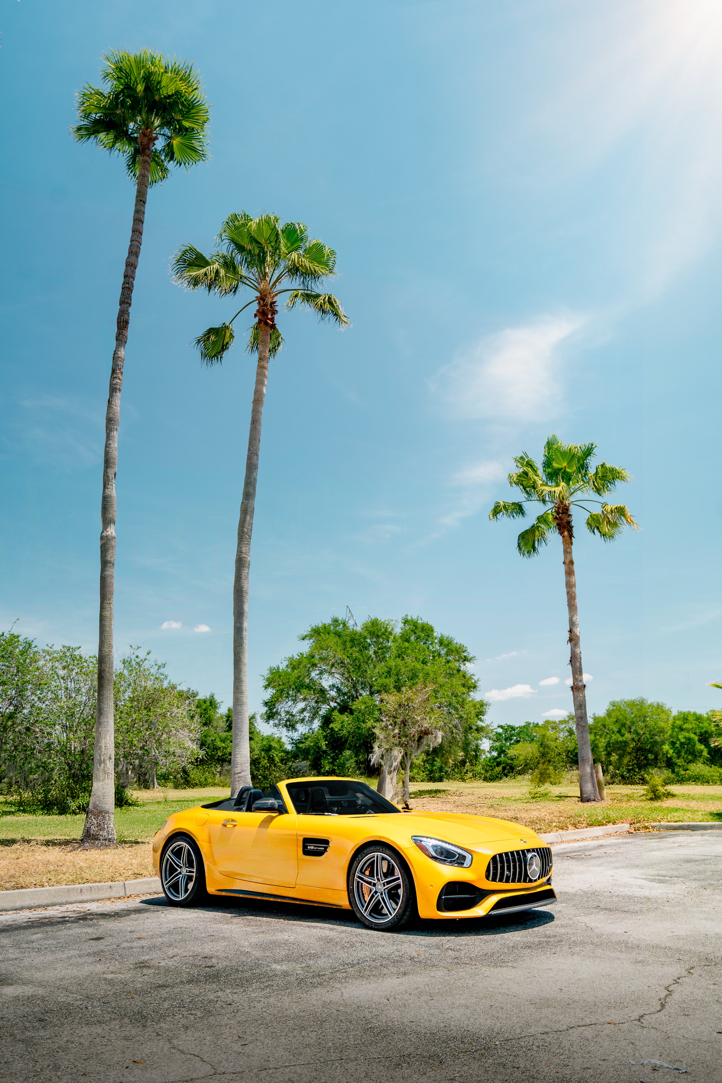 A bright yellow Mercedes-Benz convertible sports car parked on a paved lot with palm trees and green trees in the background under a clear blue sky in Orlando