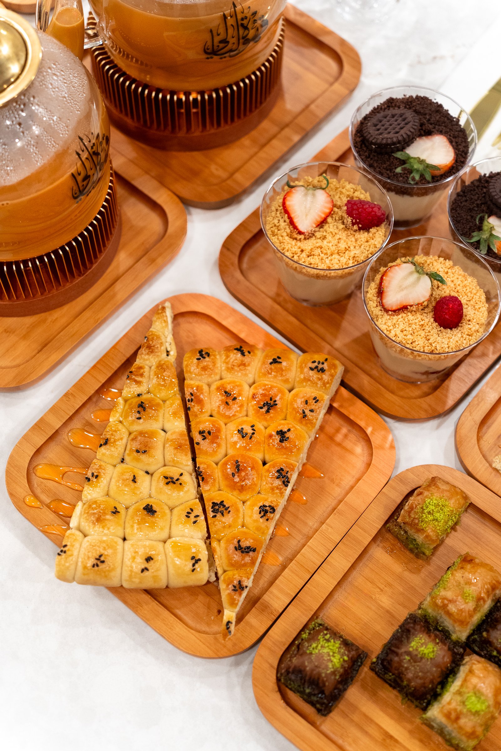 Assorted Middle Eastern desserts and drinks on wooden trays, including baklava rolls, a sliced pastry with sesame seeds, and cups of pudding topped with strawberries, raspberries, crushed cookies, and Oreos.