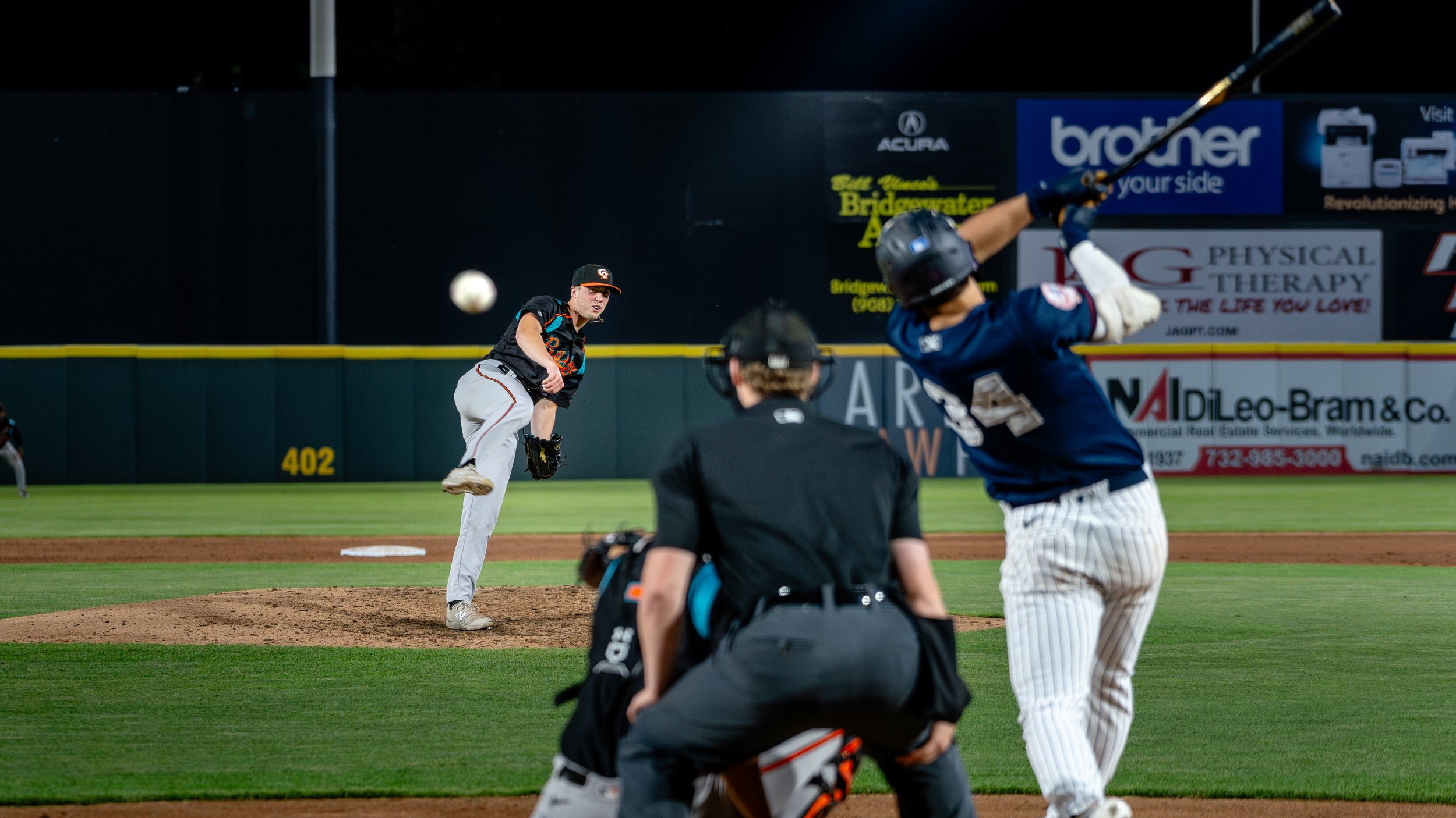 A baseball pitcher in a black cap and black jersey throws a pitch, while a batter in a navy blue jersey and white pinstriped pants prepares to swing, an umpire crouches behind them observing the play, on a baseball field at night.
