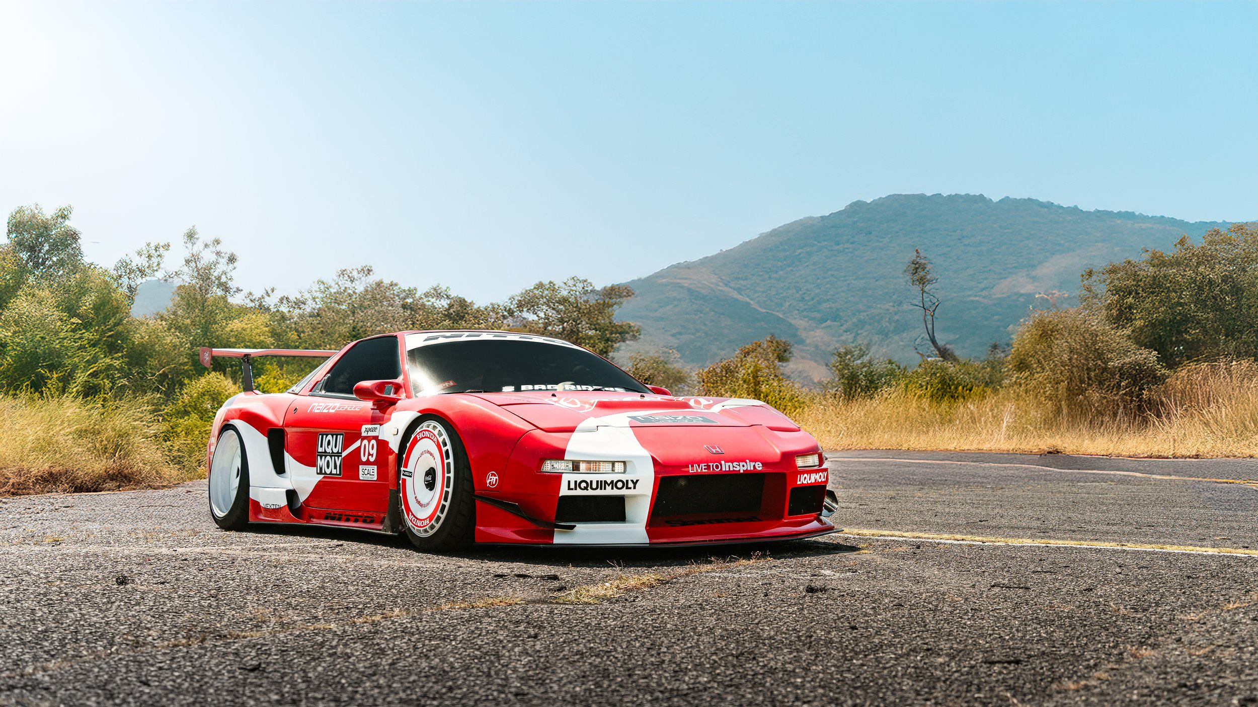 Red and white custom wrapped lowered NSX on an empty road with mountains and dry grass in the background.