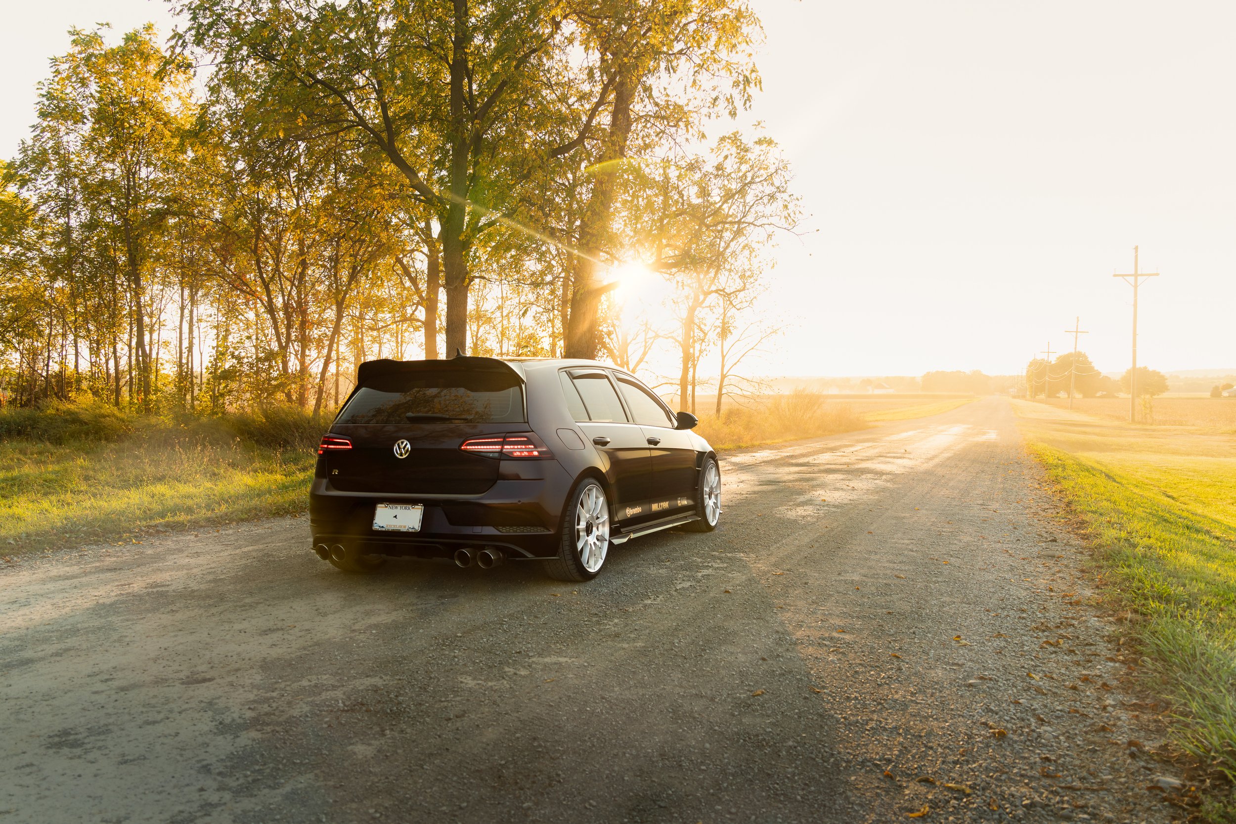 A black Volkswagen Golf R parked on a rural dirt road next to a grassy area, with tall trees and power lines in the background during sunset.