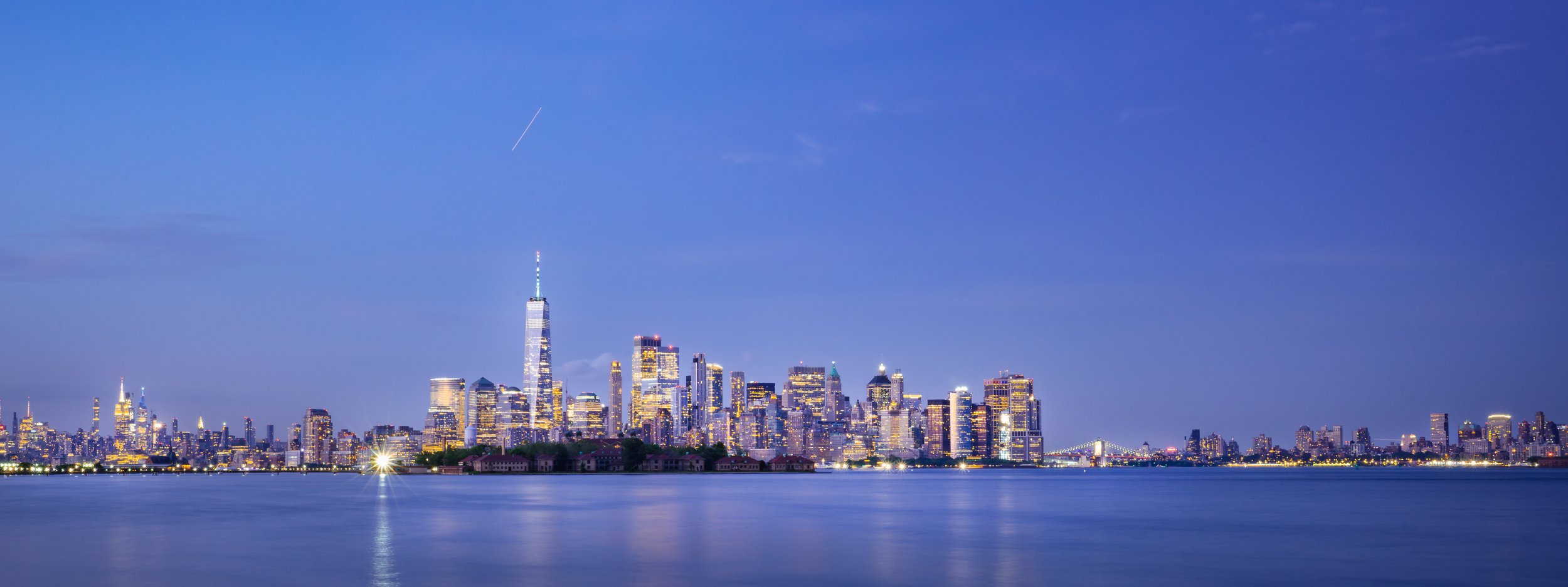 Nighttime view of the New York City skyline, including the One World Trade Center, with calm water in the foreground and a clear sky above.