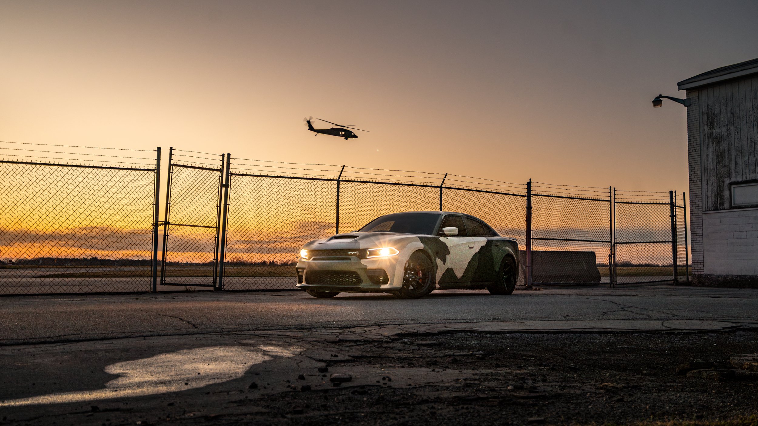 Camo Dodge Charger SRT parked behind a chain-link fence at sunset, with a black hawk military helicopter flying in the sky nearby.