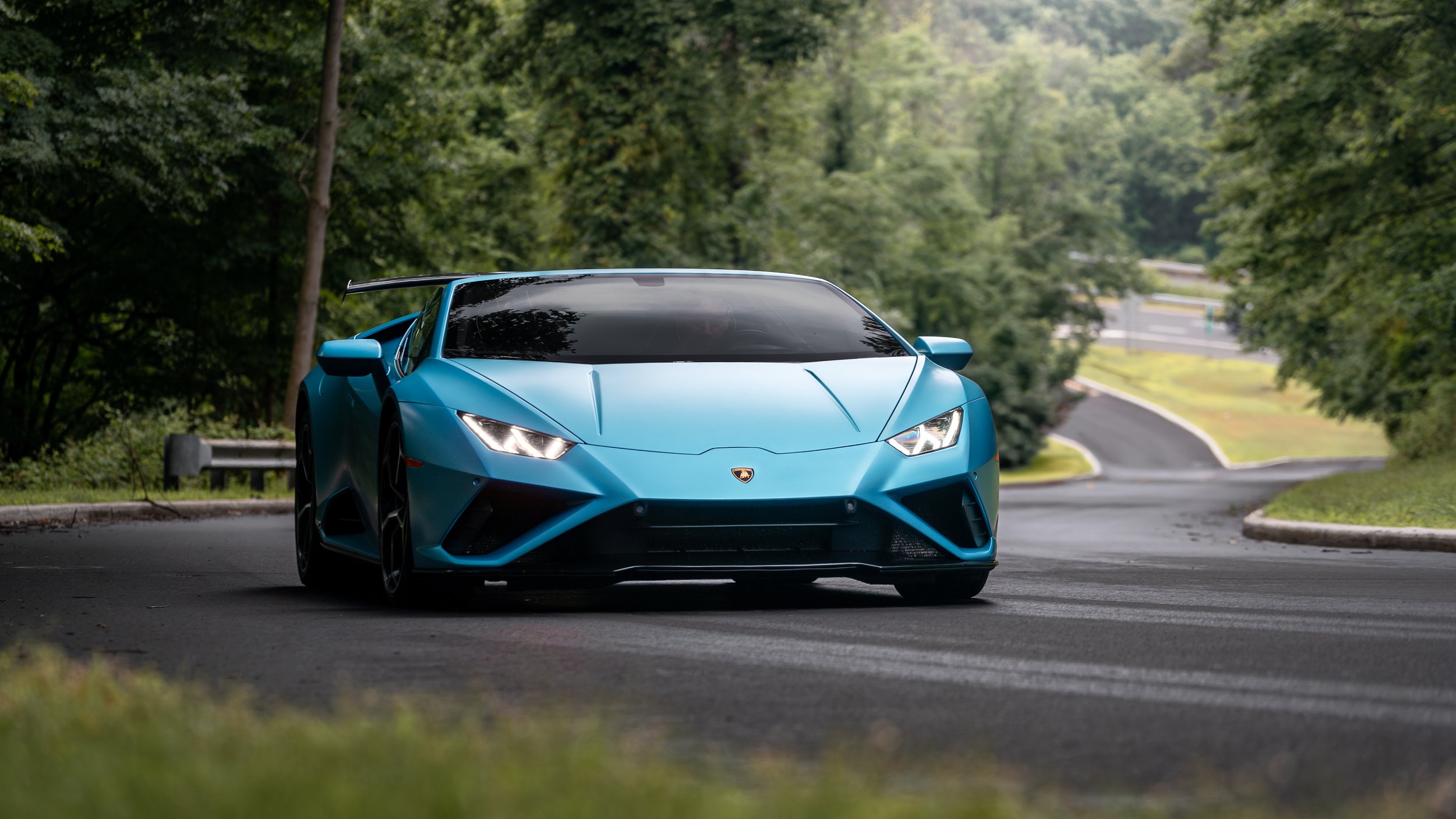 A blue Lamborghini Huracan Performante driving on a winding road surrounded by green trees.