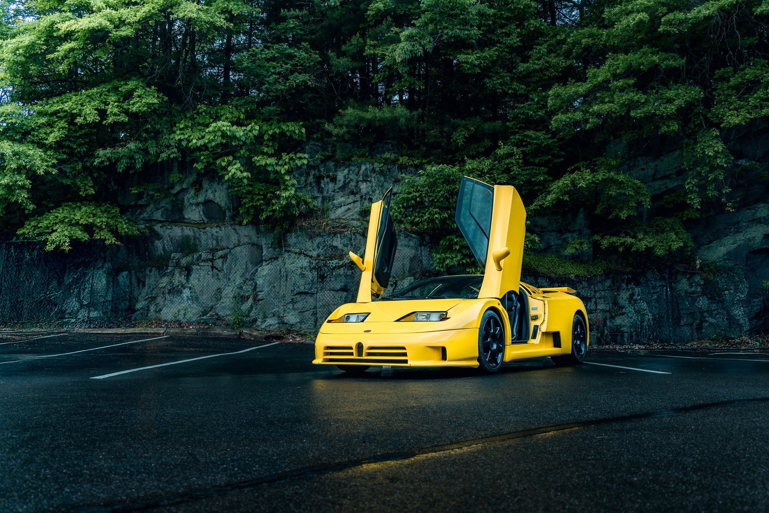 A Bugatti EB110 with vertical gull-wing doors open, parked on a wet asphalt road with a rocky and green wooded background.