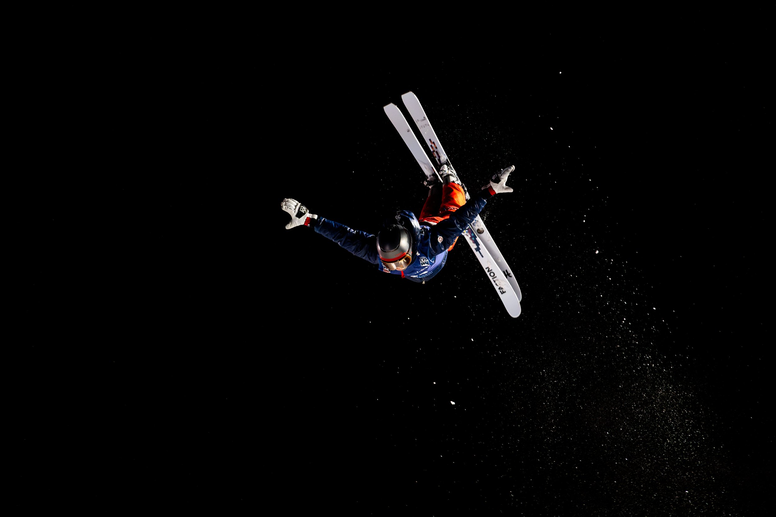 Skier wearing blue, red, and black gear executing a mid-air stunt against a black background, with snow spray at Bristol Ski Resort