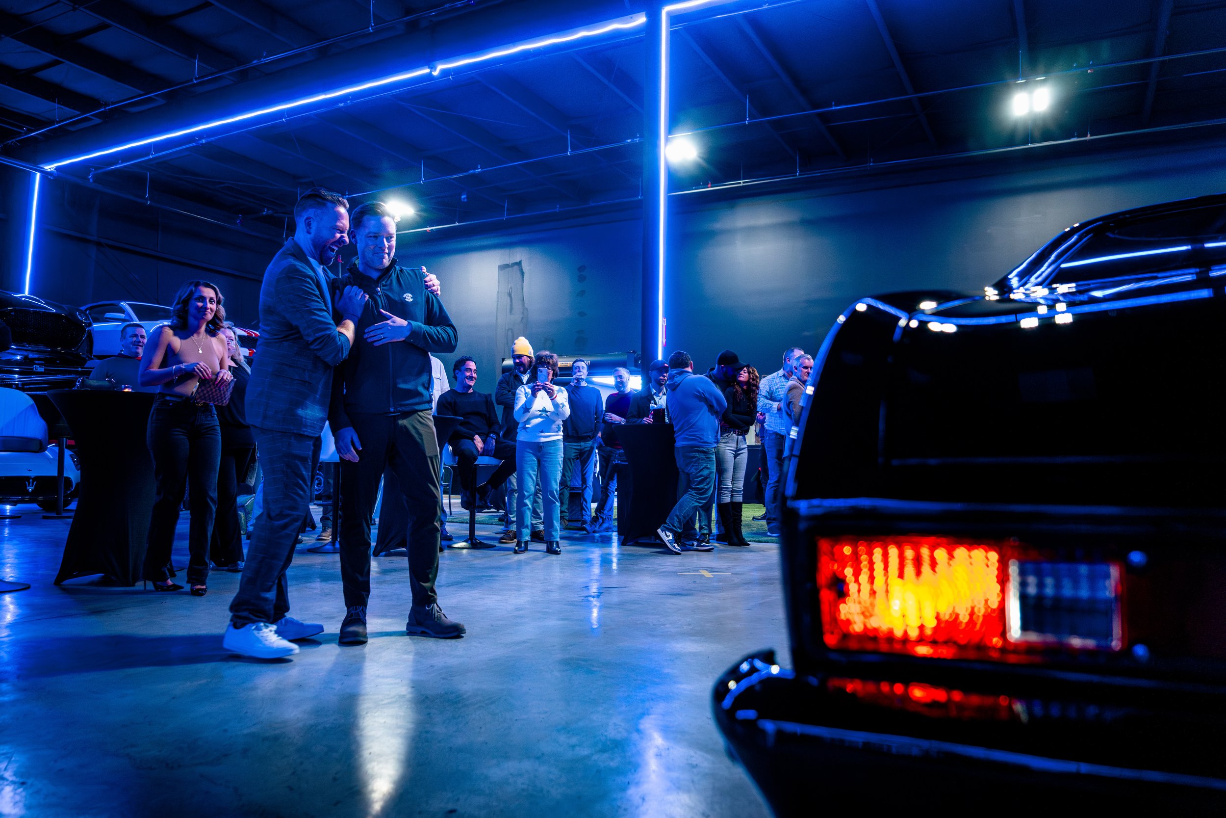 People gathered in a showroom with neon blue lighting and luxury cars, some standing and talking, others taking photos, at a social event at the Car Club of Rochester