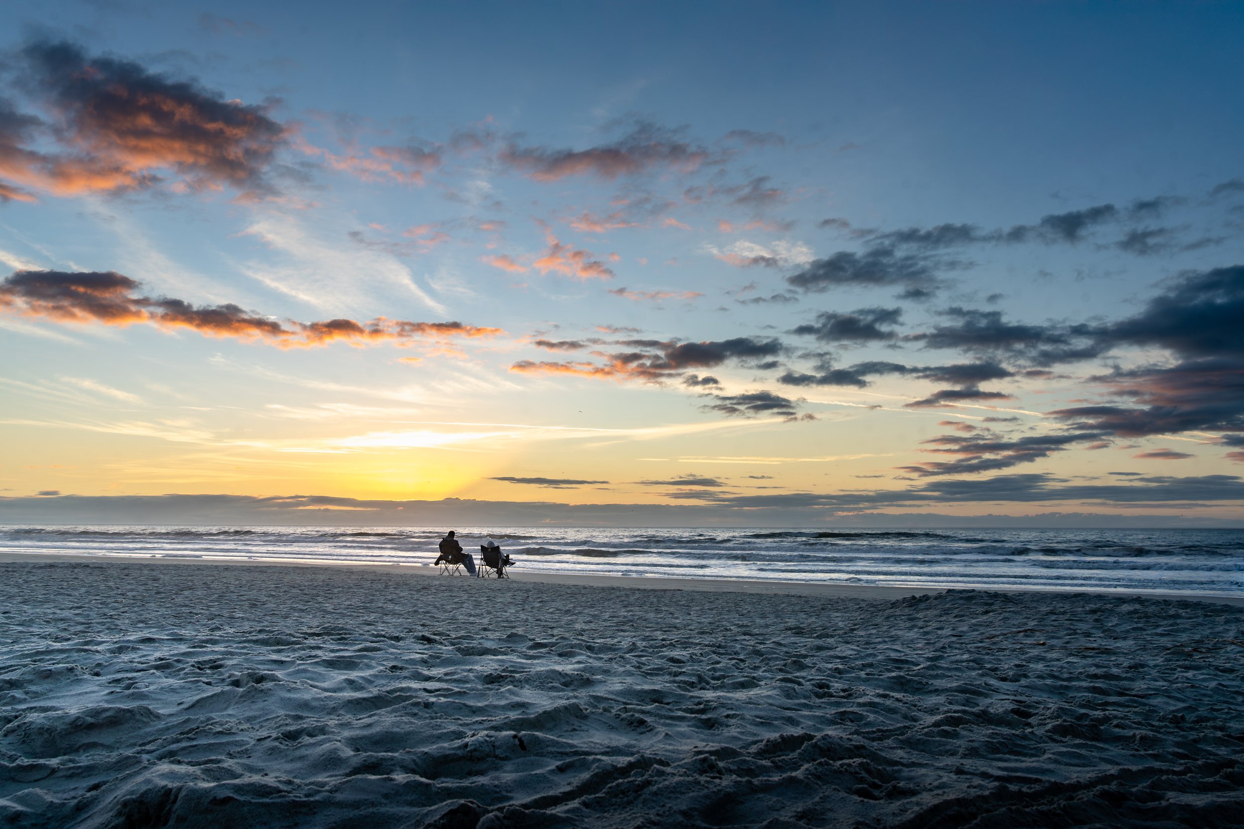 People sitting on the beach at sunset, watching the ocean and clouds.