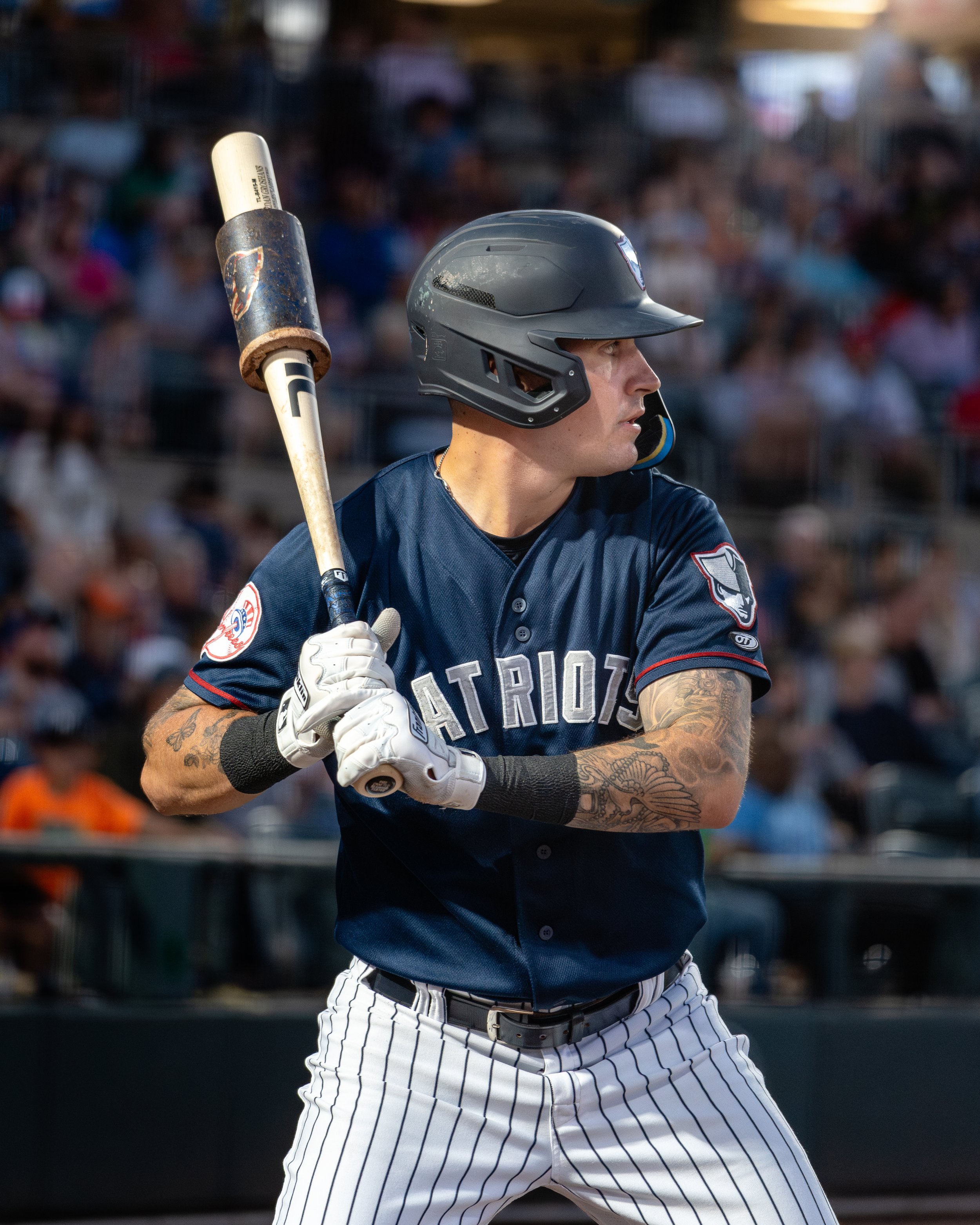 A baseball player stands at home plate, wearing a navy blue uniform with the words 'Patriots' on it, white striped pants, a black helmet, and holding a bat ready to swing, with a blurred crowd in the background.