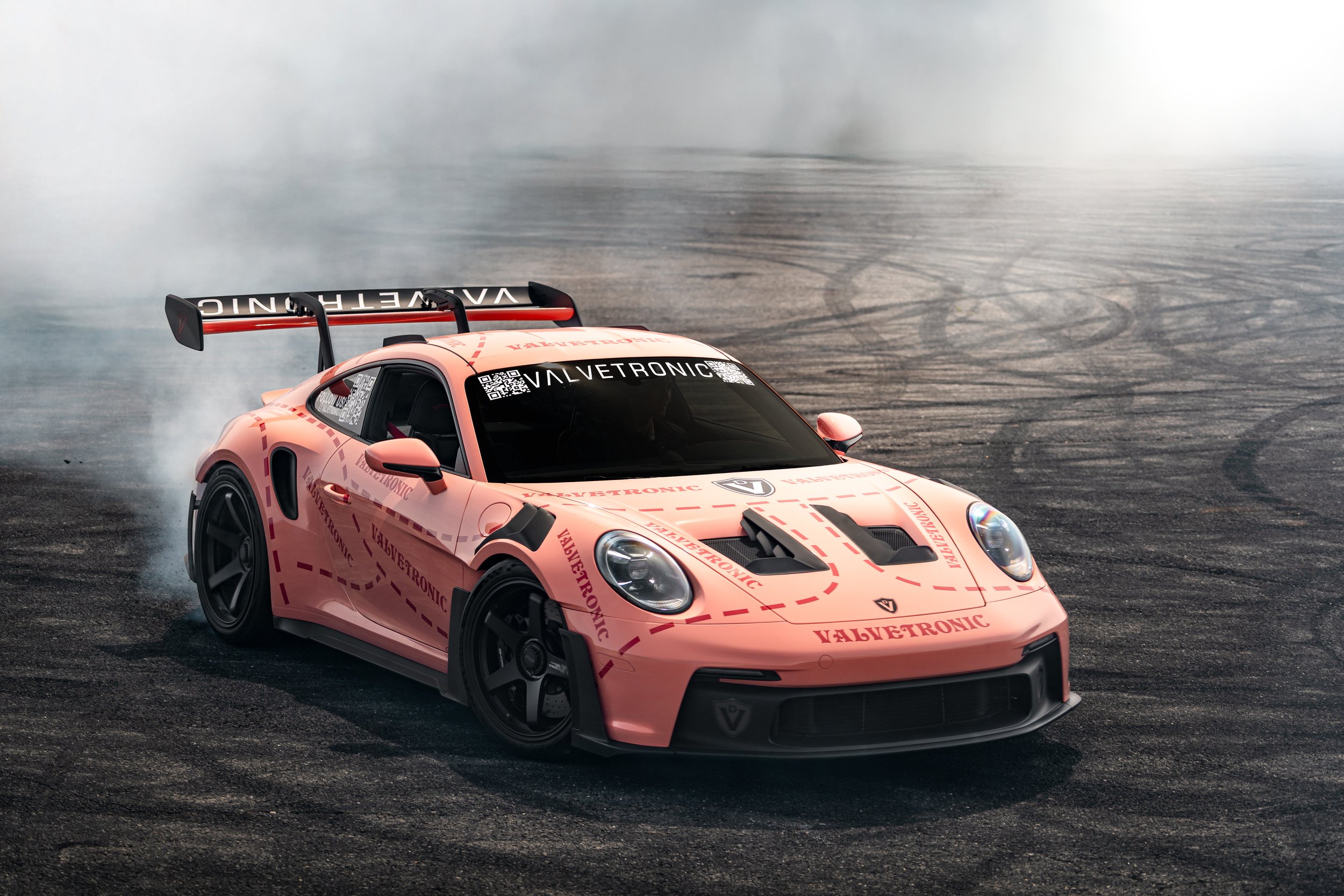 Valvetronics Porsche GT3rs drifting on a track, kicking up smoke, with VALVETRONIC branding on its hood and windshield, and a black rear wing.