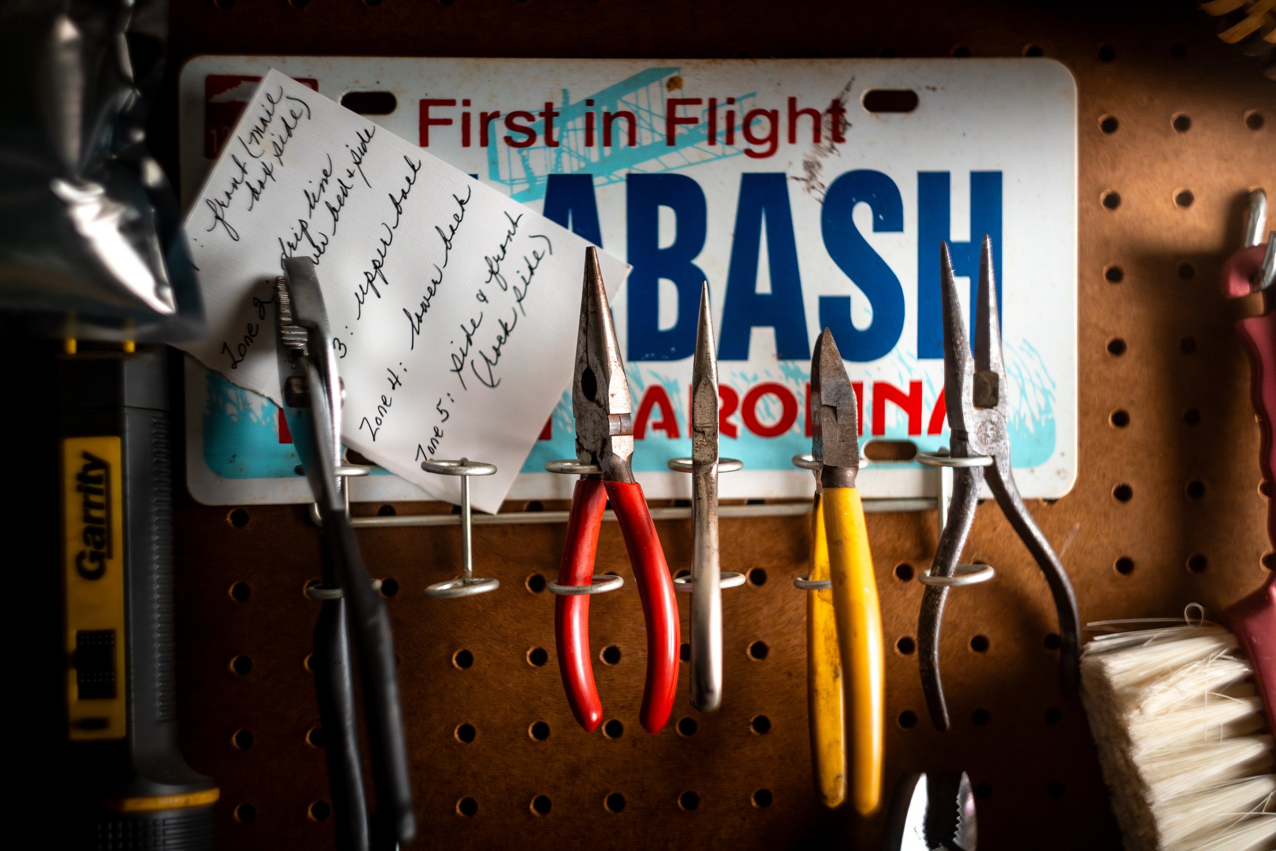 A car bumper sticker with the words 'First in Flight, Bash Arizona' behind a collection of hand tools hanging on a pegboard, including pliers, needle-nose pliers, and a brush.