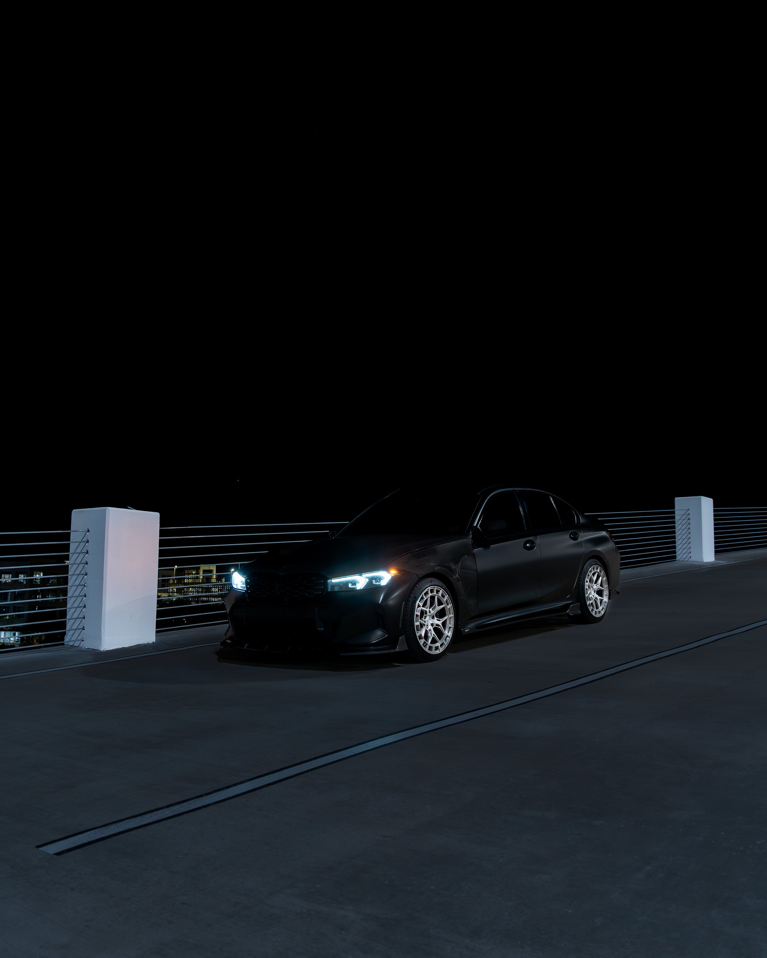 Black car with illuminated headlights parked on a rooftop at night, with city lights in the background.