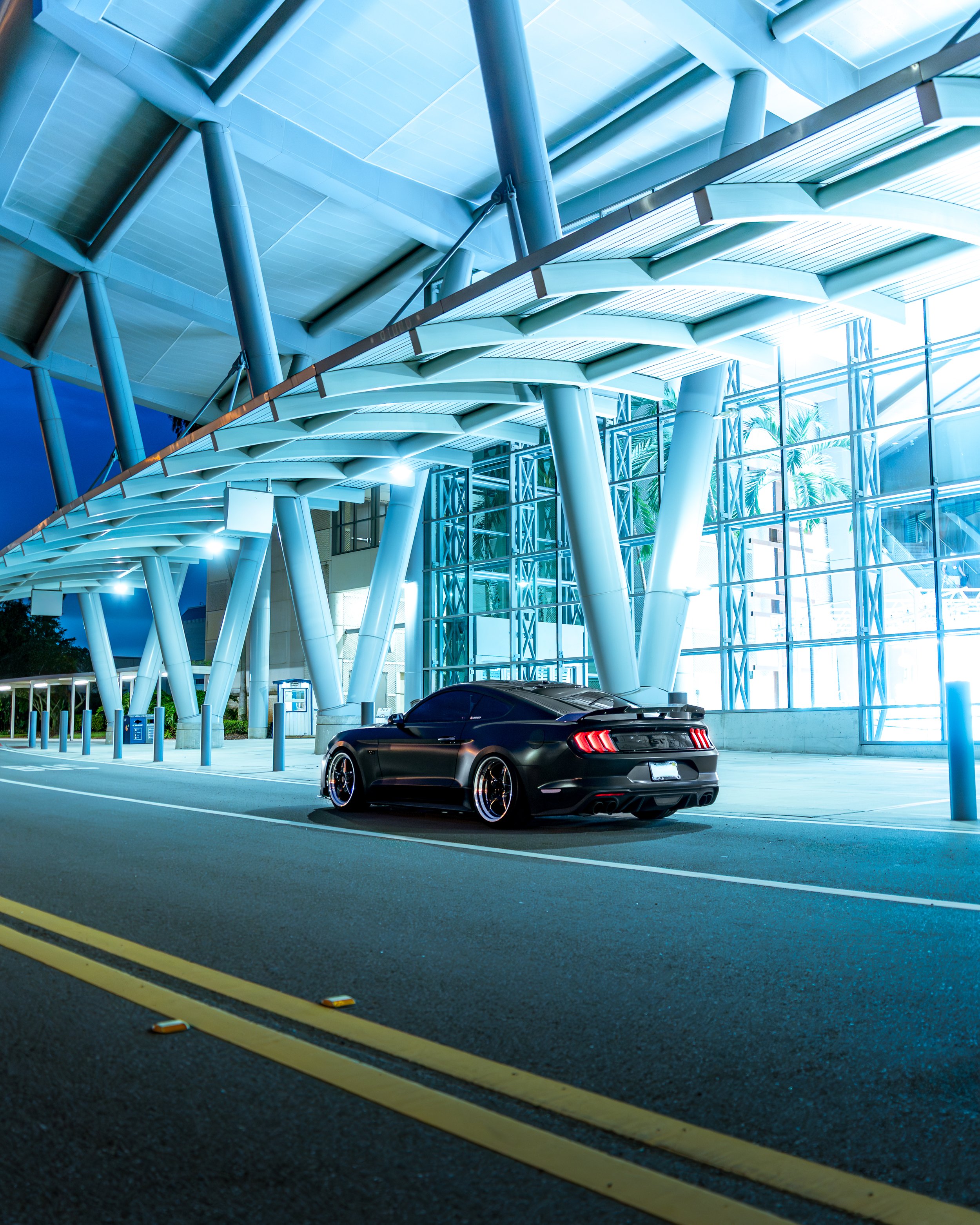Black Mustang 5.0 parked outside a modern architectural building with large glass windows and structural steel supports at dusk or nighttime.
