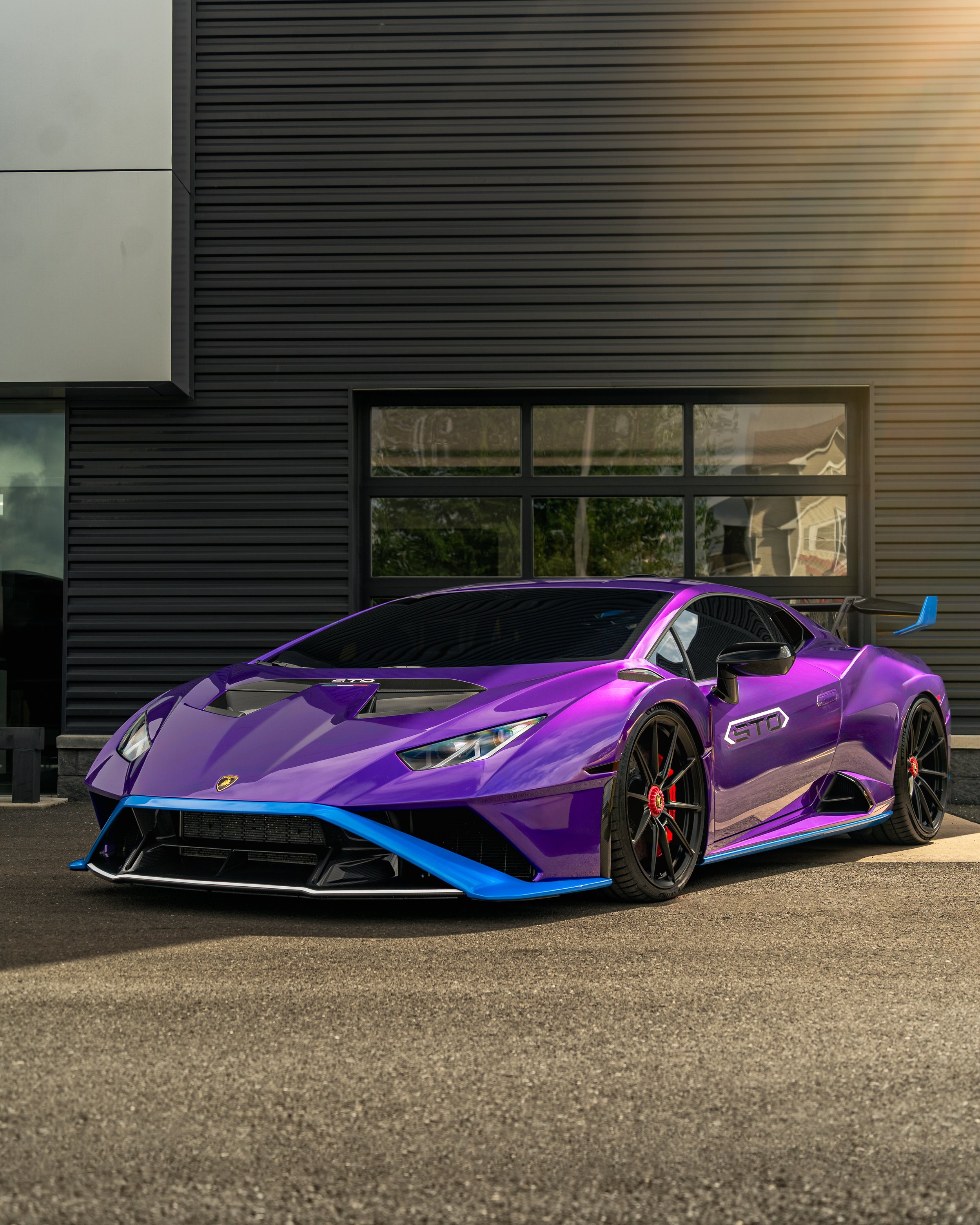 A purple Lamborghini Huracán STO parked outside a modern building with black siding and windows, featuring a blue front splitter, black wheels with red brake calipers, and a rear wing.