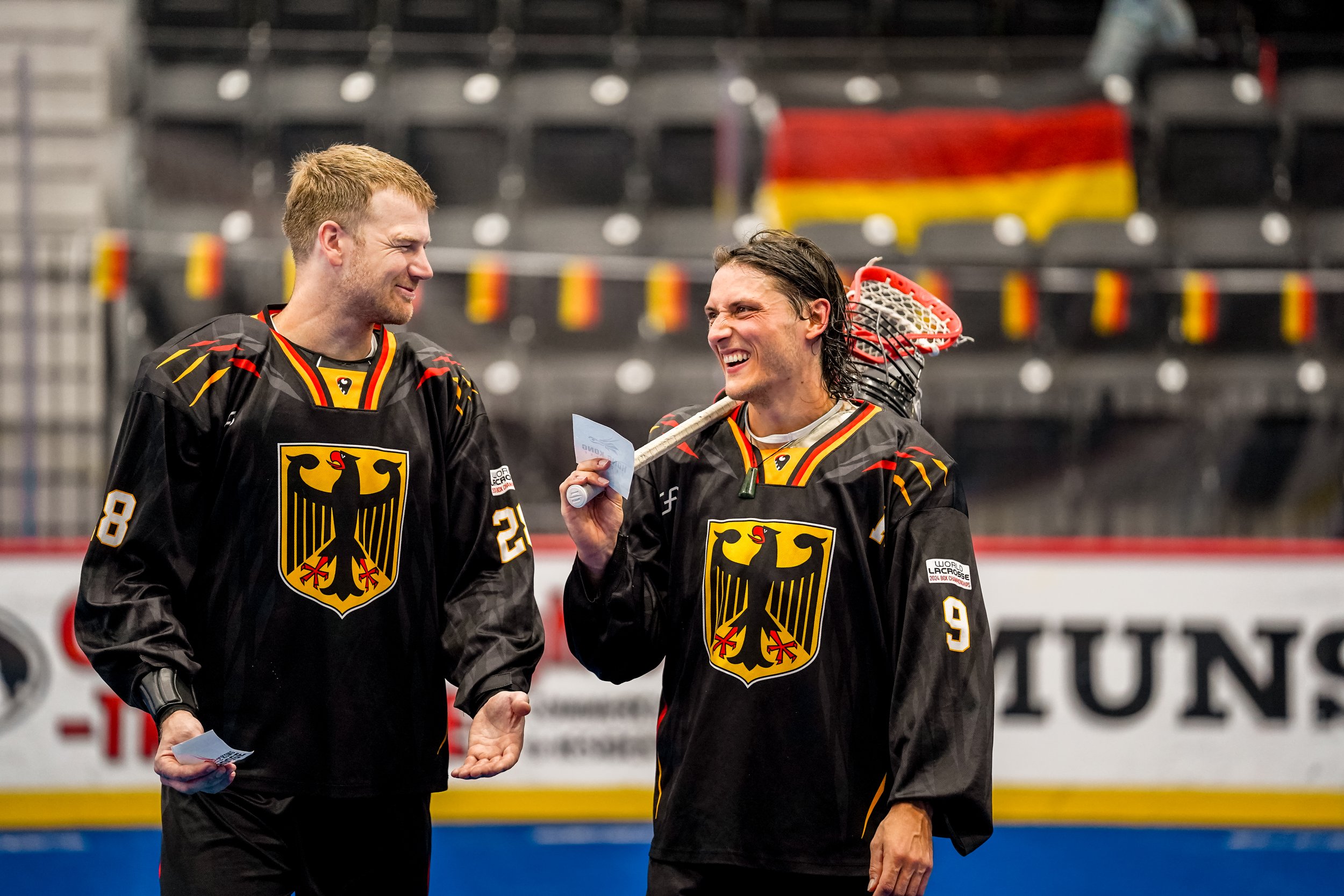 Two male ice hockey players in black jerseys with the German eagle emblem, smiling and talking on the ice rink, one holding a hockey stick over his shoulder.