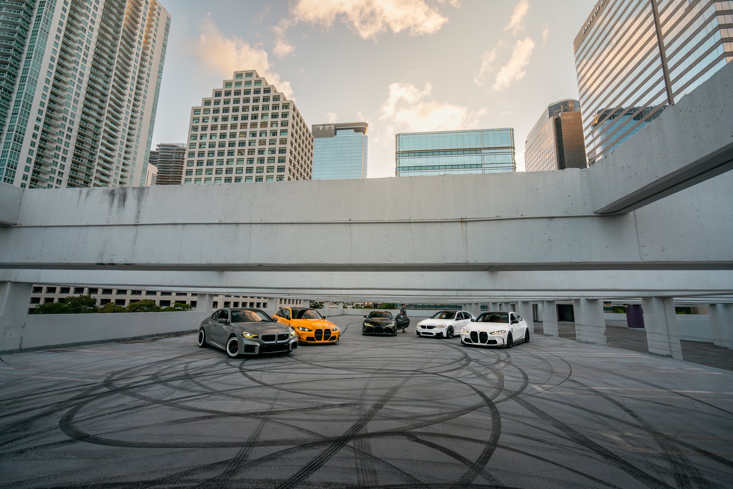 Five luxury sports cars parked on a rooftop parking garage with a city skyline of tall buildings in the background in Miami