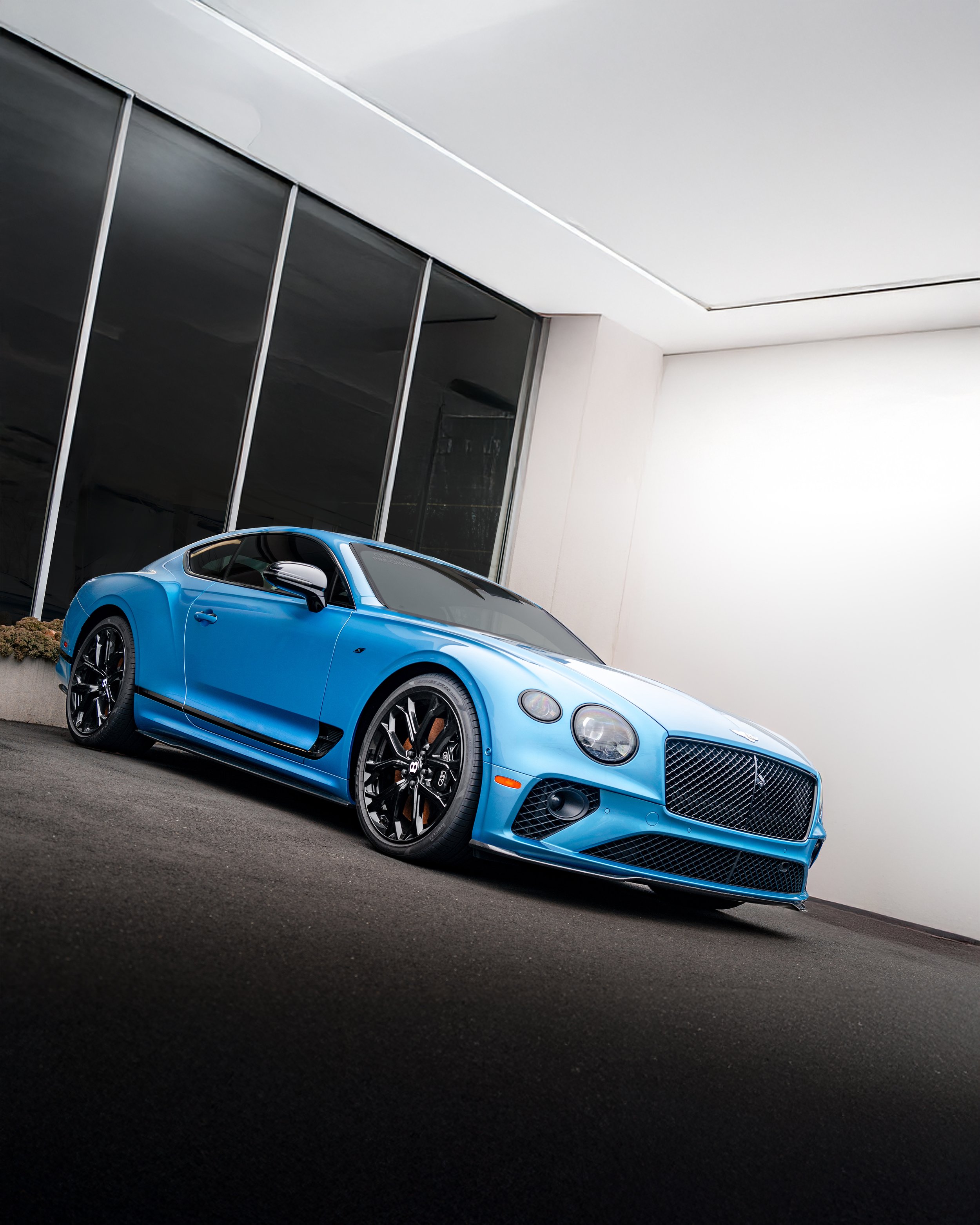 A blue Bentley Continental GT parked outside a modern building with large glass windows.