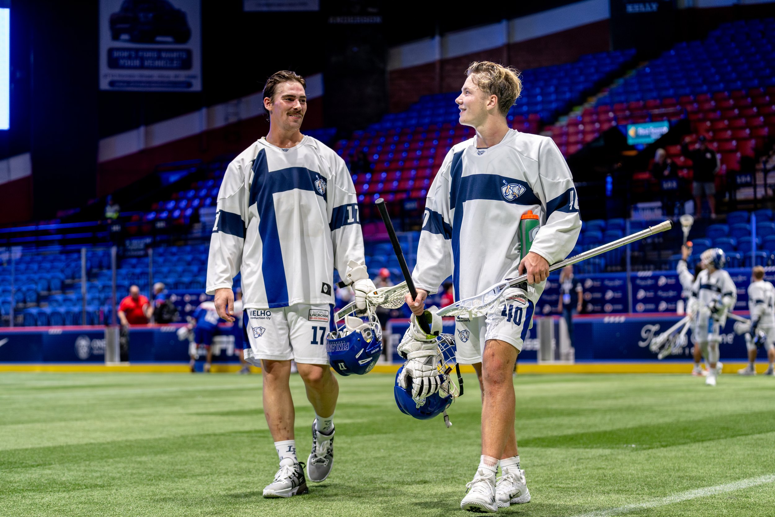 Two male lacrosse players from Team Finland walking and talking on a field inside a stadium, holding lacrosse sticks and helmets at the World Box Lacrosse Tournament in Utica, NY