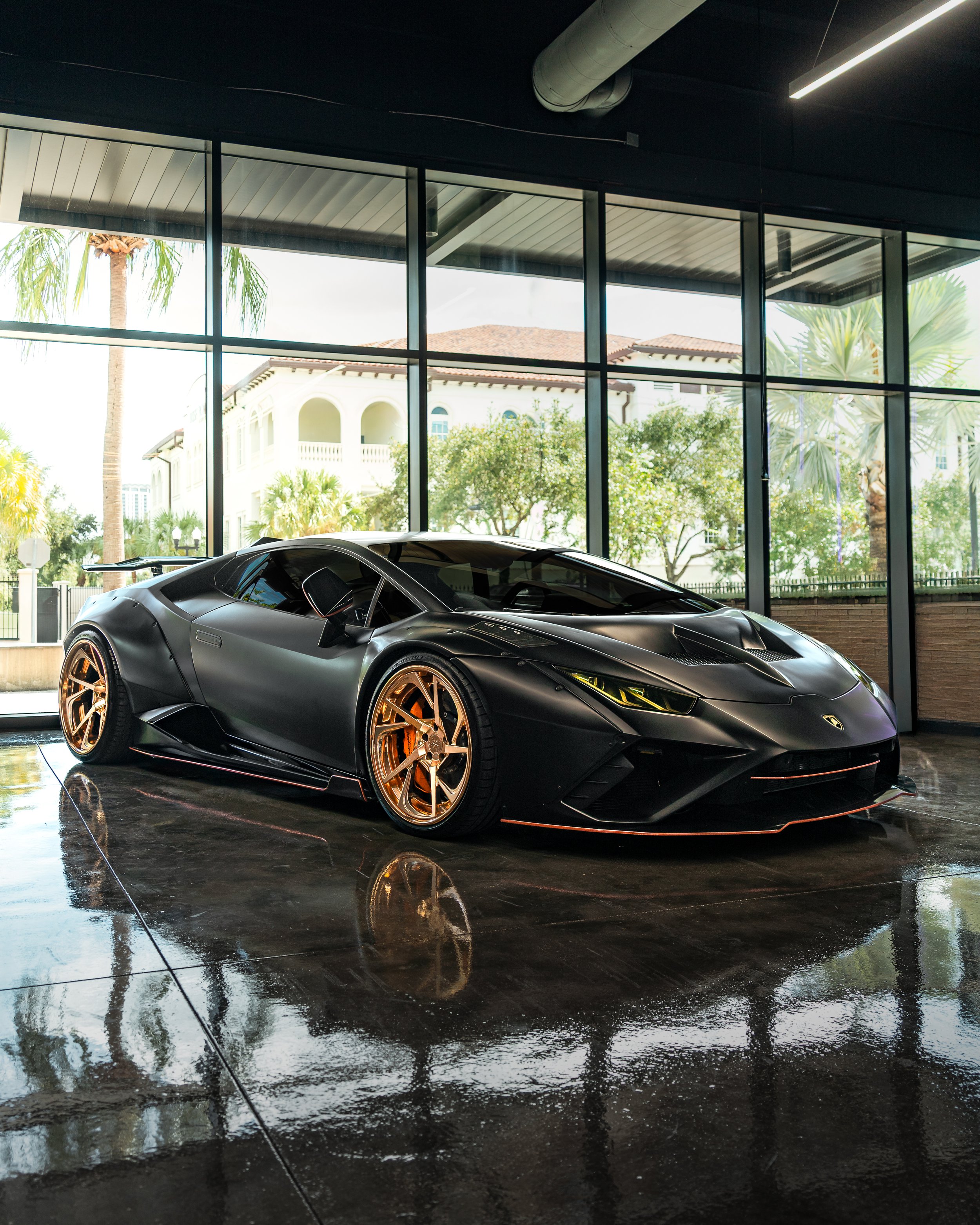 A black Liberty Walk widebody one of one Lamborghini Huracan Performante sports car with gold wheels parked indoors near large glass windows showing palm trees and residential buildings outside.