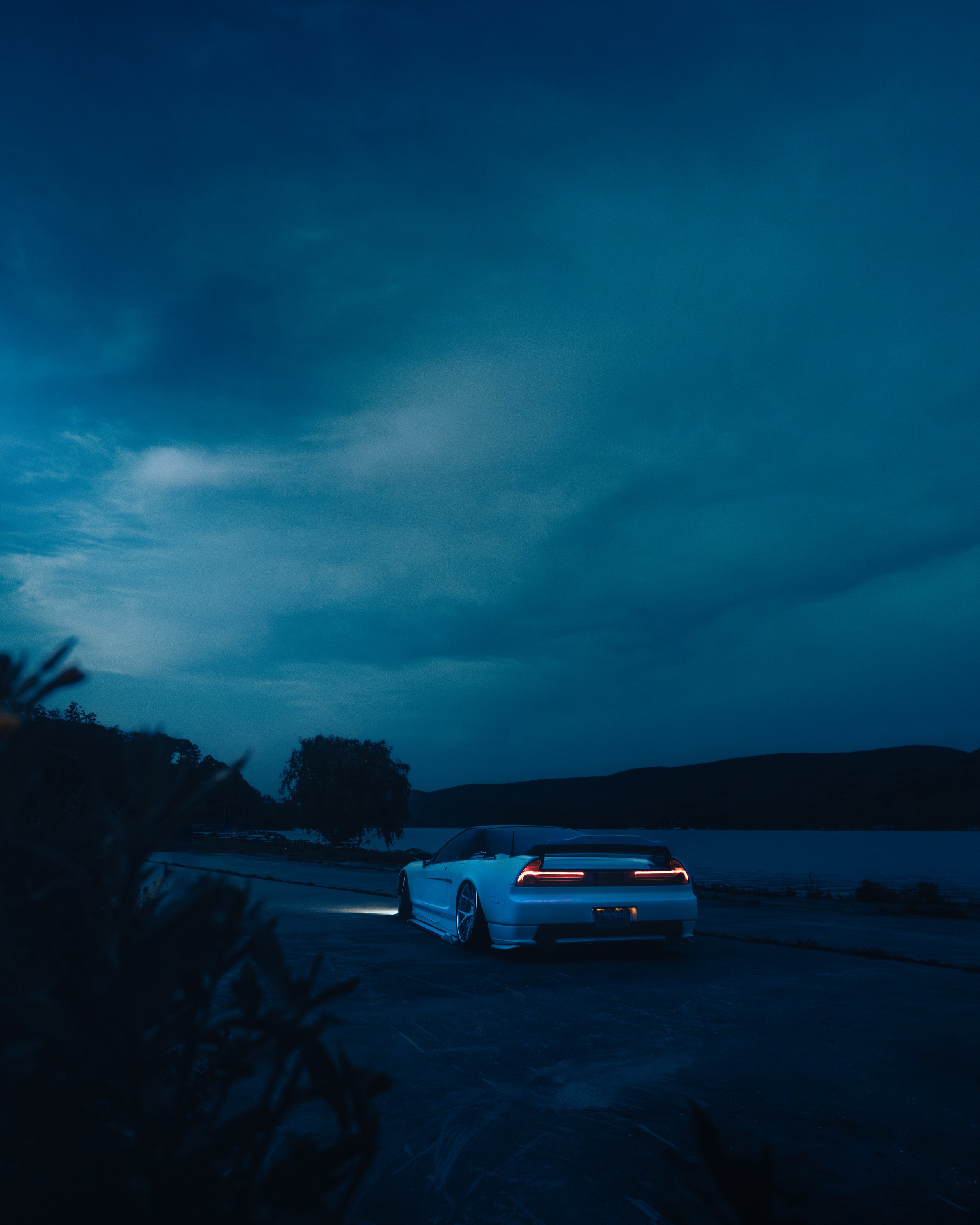 A lowered Honda NSX with glowing taillights parked near a body of water under a dark, cloudy sky at dusk or dawn.